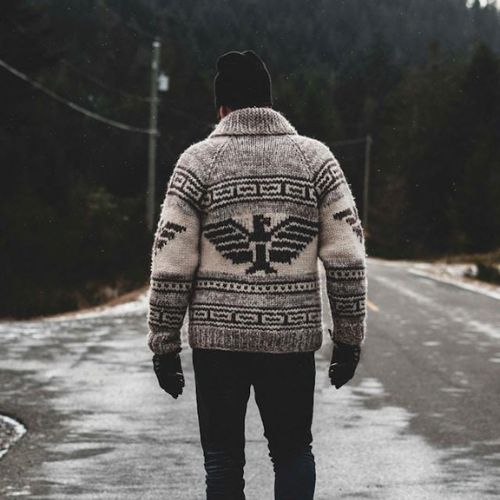 A man walking away from the camera, wearing a patterned sweater, black jeans, and boots, with snow lightly falling in a wintry, forested landscape.