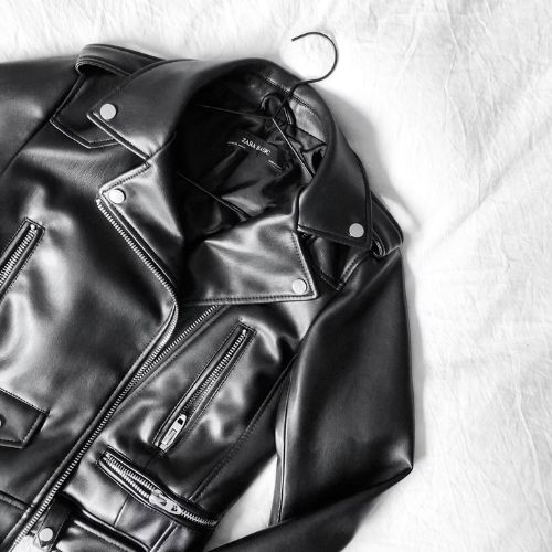 A black leather jacket on a white background, featuring multiple zippers and a glossy finish, displayed on a coat hanger to emphasize its sleek design.