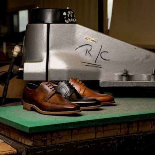 A pair of polished brown leather oxford shoes placed on a green mat near a heavy-duty shoe press in an industrial setting.