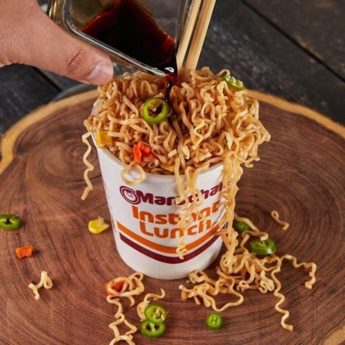 A hand pouring a dark sauce from a small glass pitcher over a cup of instant noodles. The noodles are overflowing from the cup labeled "Maruchan Instant Lunch," with chopped green chili peppers and other vegetables scattered around