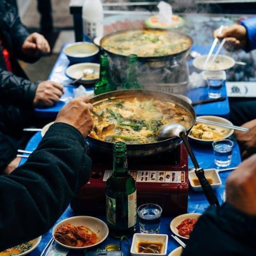 A table full of dishes and food items at a bustling restaurant. Prominently in the foreground, there is a hot pot with boiling soup and ingredients like vegetables and meat