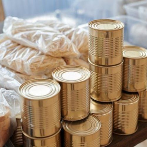 A stack of shiny, gold-colored tin cans arranged in a pyramid shape on a wooden table. Surrounding the cans, there are plastic bags filled with various groceries, such as pasta and bread, neatly organized