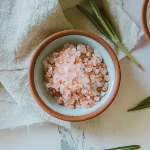 A small bowl filled with coarse pink Himalayan salt displayed on a light-colored fabric napkin. Next to the bowl, there are fresh rosemary sprigs and a cup of yellow olive oil
