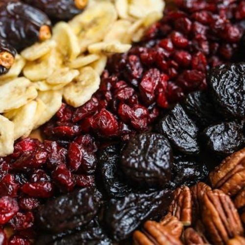 A close-up assortment of dried fruits and nuts displayed in neat, separated rows. The selection includes dried bananas, cranberries, prunes, dates, and pecans