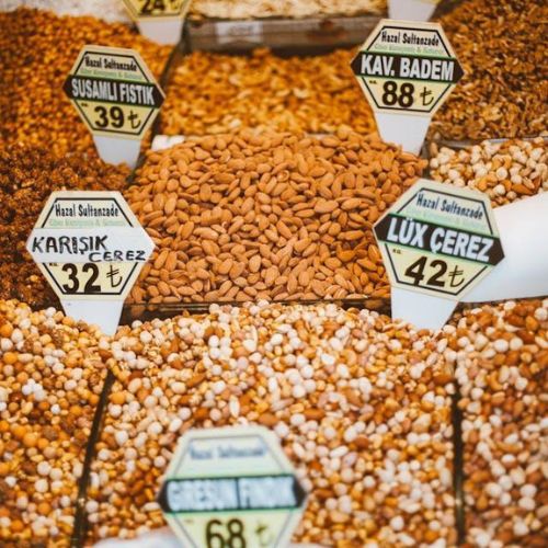 Various types of nuts and dried fruits are presented in an indoor market. Each pile of nuts, such as almonds, peanuts, and cashews, has a sign with prices listed in Turkish lira