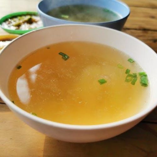A close-up view of a white bowl filled with clear, golden broth garnished with chopped green onions, placed on a wooden table