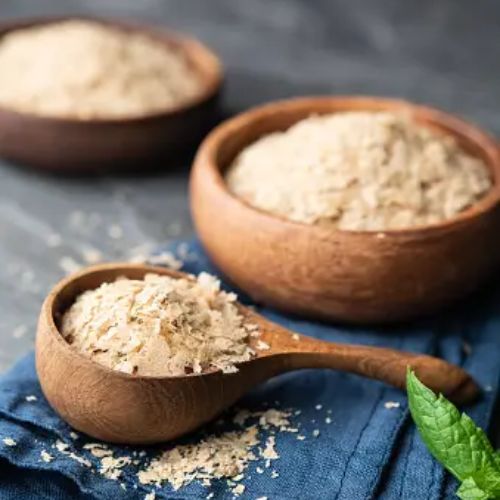 Two wooden bowls filled with nutritional yeast flakes, one with a wooden spoon, set on a navy blue cloth over a rustic table.