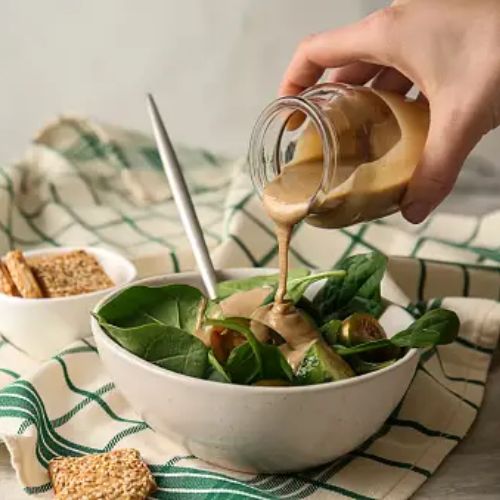 A hand pouring tahini over a salad in a ceramic bowl, surrounded by ingredients and condiments on a kitchen towel.