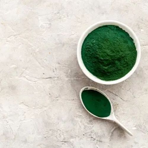 A bowl of spirulina powder next to a spoonful of the algae-based supplement, set on a gray textured background.