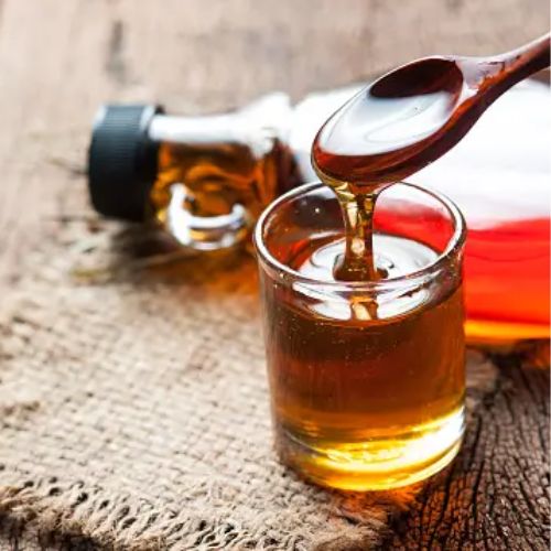 Maple syrup being poured from a wooden dipper into a small glass, with a full bottle of maple syrup next to it on a rustic wooden surface.