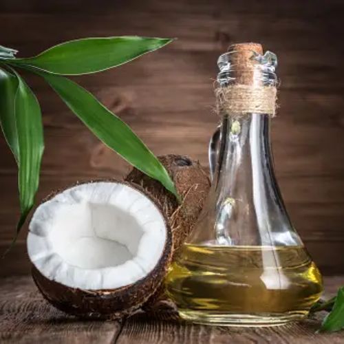 A clear glass bottle of olive oil with a cork stopper, alongside a halved coconut and green leaves, placed on a wooden surface.