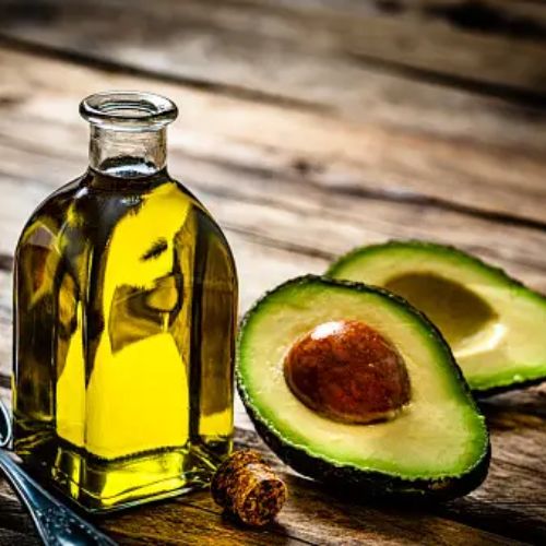 Avocado halves with a bottle of avocado oil and a spoonful of oil, displayed on a rustic wooden table.