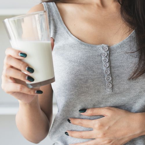 A woman holding a glass of milk, with her other hand on her stomach, suggesting she may be experiencing discomfort or focusing on digestive health. She is wearing a grey tank top with decorative buttons.