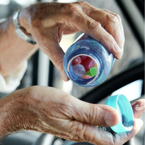 Elderly hands holding and opening a small blue bottle containing colorful candies, depicting possibly a need or a habit of taking regular medication.