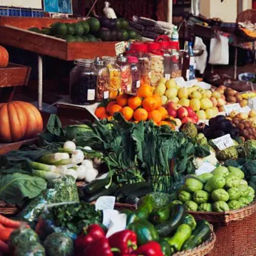 A vibrant and abundant display of fresh vegetables and fruits at a market stand, including greens, oranges, pumpkins, and assorted produce with jars of spices in the background.