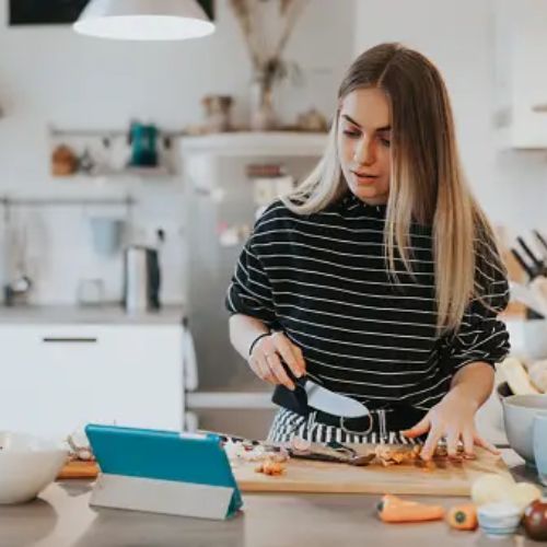 A woman in a black and white striped top is intently chopping vegetables on a wooden cutting board in a modern kitchen, with a tablet displaying a recipe propped up in front of her.