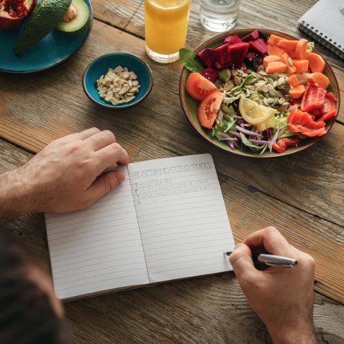 An overhead view of a person writing a meal plan in a notebook, with a variety of colorful fruits and salads on the table.