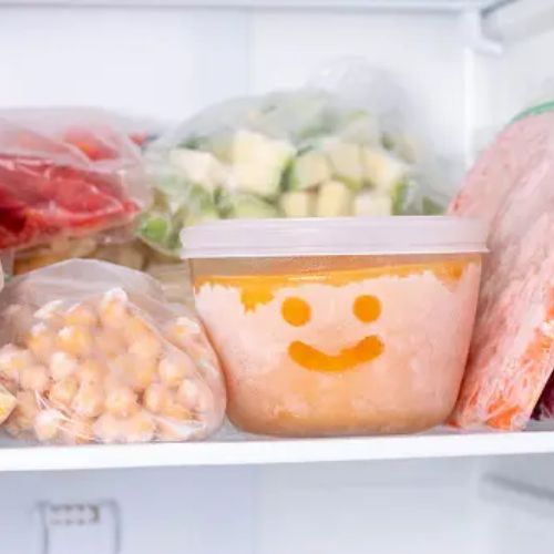 A playful container of chilled pureed food with a smiley face drawn in the condensation, surrounded by other frozen food items in a fridge.