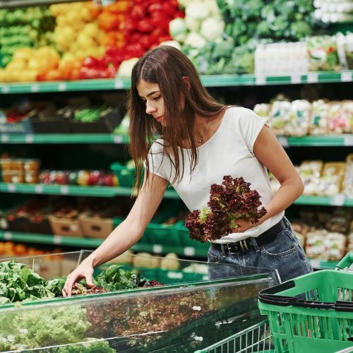 A woman selecting fresh lettuce from a grocery store produce section, placing it into a green shopping basket.