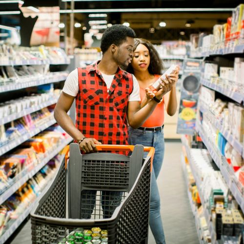 A young couple shopping in a supermarket, the man pushing a cart while the woman is showing him a product.