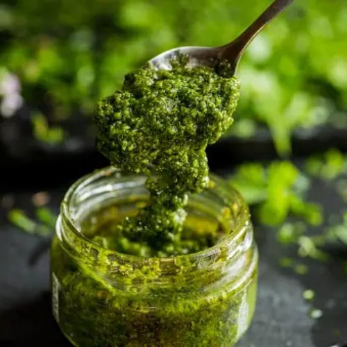 A glass jar of fresh homemade green pesto sauce, with a spoon scooping it out, set against a lush green, plant-filled background.