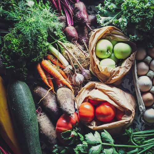 An array of fresh vegetables and fruits, including beets, kale, carrots, green apples, tomatoes, eggs, and herbs, artistically arranged on a floral patterned background.