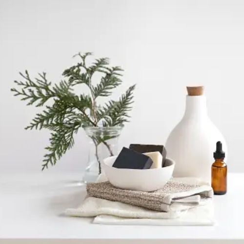 A minimalist arrangement on a white surface, featuring a bowl with various soaps, a brown bottle, stacked towels, and a vase with green branches