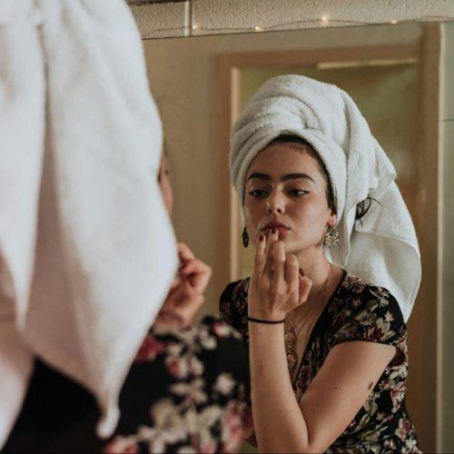 A woman with a towel wrapped around their head, applying makeup or skincare in front of a mirror