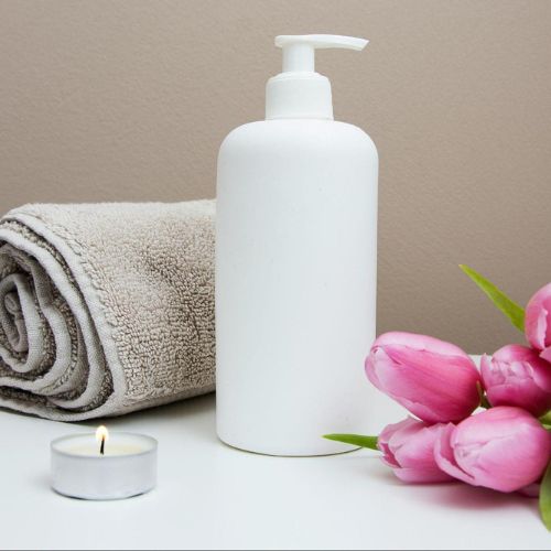 A rolled towel, a white pump bottle, a lit candle, and a bouquet of pink tulips arranged on a white surface