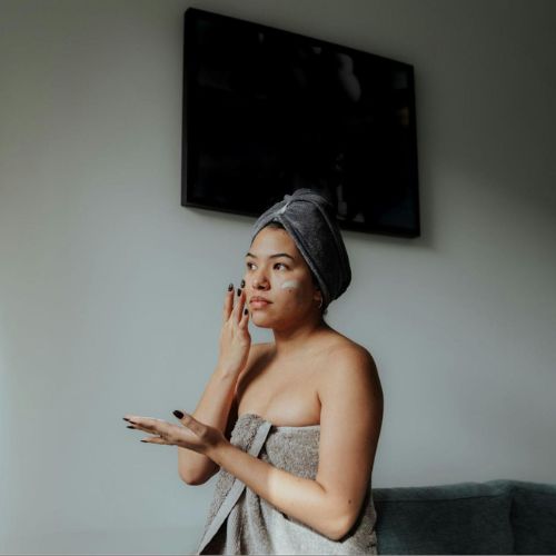 A woman wrapped in a towel with a towel on their head, applying skincare on their face in a softly lit room