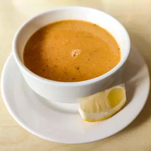 A white bowl of lentil soup on a matching white plate, served with a lemon wedge