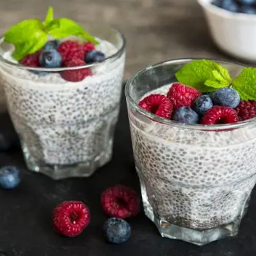 Two glasses of chia pudding topped with raspberries, blueberries, and mint leaves, with additional berries scattered around