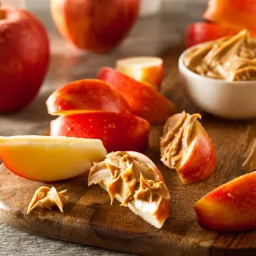 Sliced red apples with dollops of peanut butter, presented on a wooden board with whole apples and a small bowl of peanut butter in the background