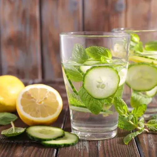 Two glasses of water infused with cucumber, lemon, and mint, placed on a wooden table with a sliced lemon and cucumber next to them