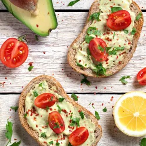 Two slices of avocado toast topped with mashed avocado, cherry tomatoes, and herbs, placed on a white wooden table with an avocado half and lemon wedge nearby