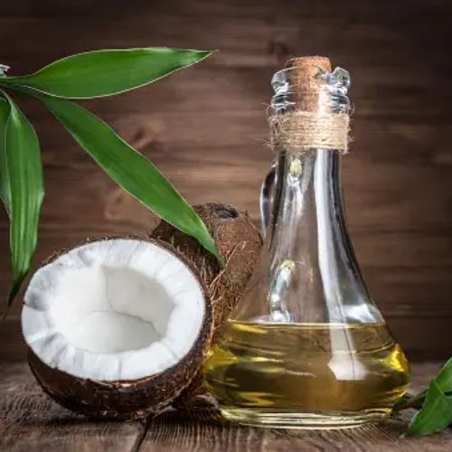 A glass bottle of coconut oil next to a halved coconut and green leaves on a wooden surface