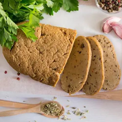 Slices of seitan on a cutting board with herbs, garlic, and a knife in the background