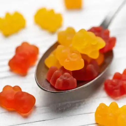 A spoon with gummy bears of different colors scattered around it on a white wooden surface