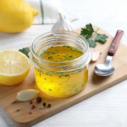 A jar of lemon herb vinaigrette with ingredients like lemon, parsley, and garlic on a wooden board