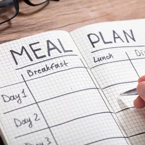 A person writing in a notebook with a meal plan layout, including sections for breakfast, lunch, and dinner for several days