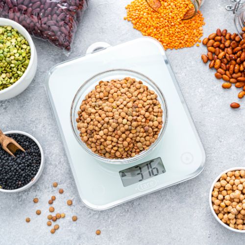 Various legumes, including lentils, kidney beans, and chickpeas, being weighed on a digital kitchen scale with more legumes scattered around on a white surface