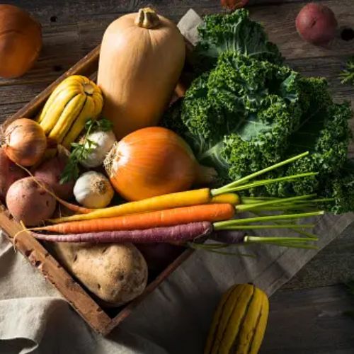 A wooden crate filled with a variety of fresh vegetables, including carrots, kale, squash, onions, and potatoes, on a rustic wooden table