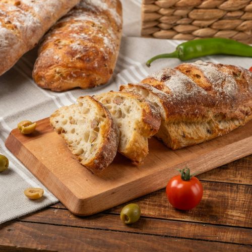 Freshly baked bread loaves on a wooden cutting board, with slices cut and ready to serve. The scene includes olives, a small tomato, and a green chili on a wooden table, creating a cozy and rustic presentation.