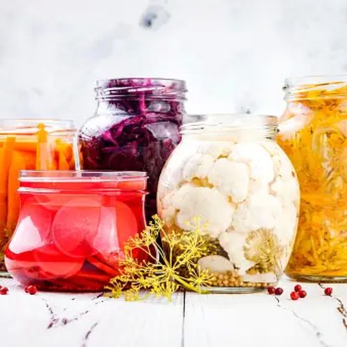 A variety of pickled vegetables in jars, including cauliflower, carrots, red cabbage, and radishes, placed on a white surface with dill and pink peppercorns scattered around.