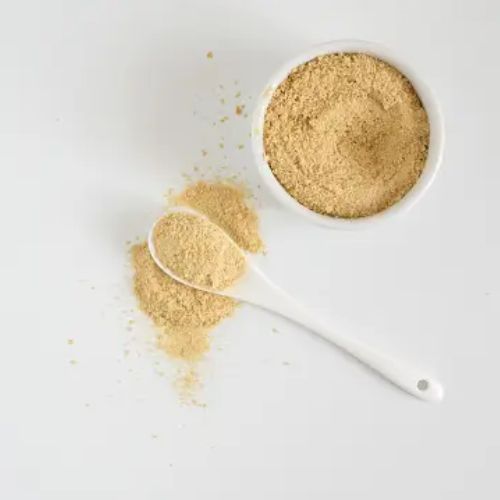 A bowl of nutritional yeast with a white spoon filled with some yeast flakes placed on a white surface