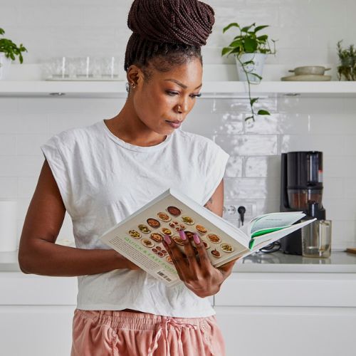 A woman standing in a kitchen, wearing a white t-shirt and pink pants, deeply focused on reading a cookbook. There are plants and kitchen appliances in the background.