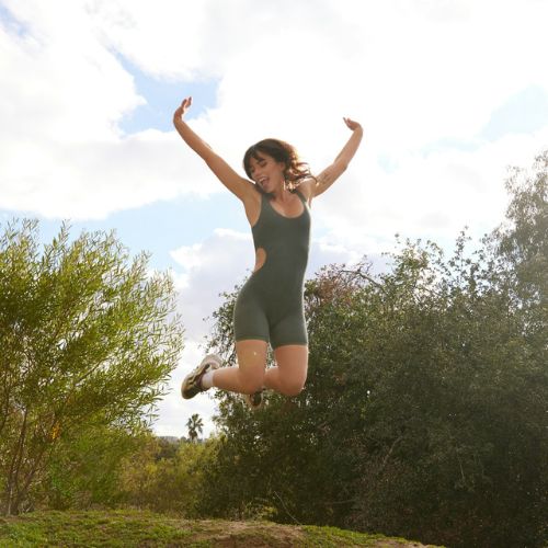 A woman in a sporty outfit, jumping joyfully in the air with her arms raised against a backdrop of trees and a partly cloudy sky.