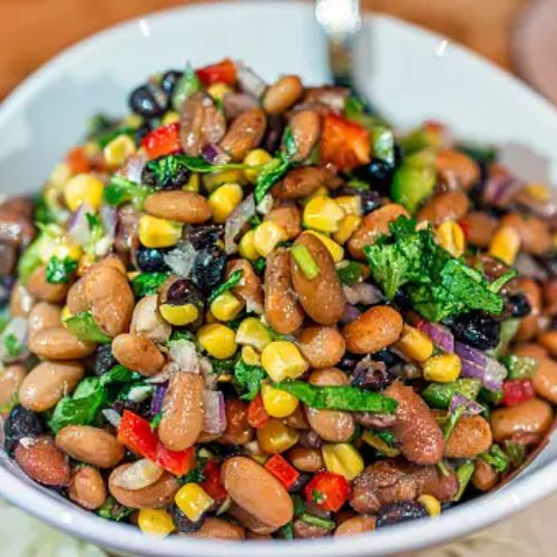 A colorful bean salad with different types of beans, corn, red peppers, and cilantro, in a white bowl