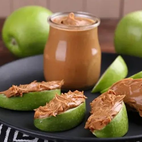 Slices of green apple spread with peanut butter, served on a black plate with a jar of peanut butter and whole green apples in the background