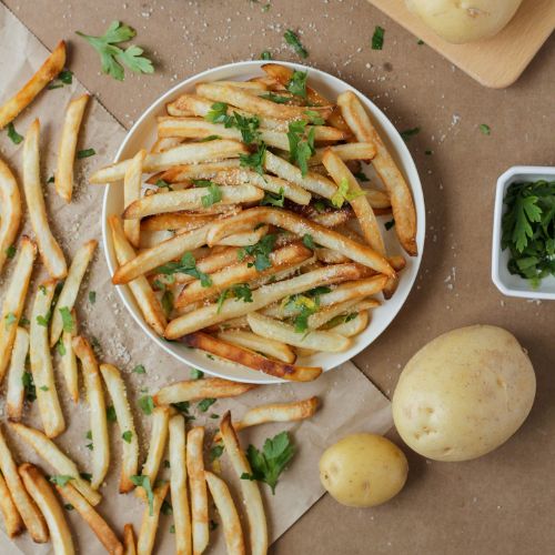 A top view of freshly cooked French fries served on a plate and sprinkled with herbs. Additional herbs, potatoes, and scattered fries surround the main plate.